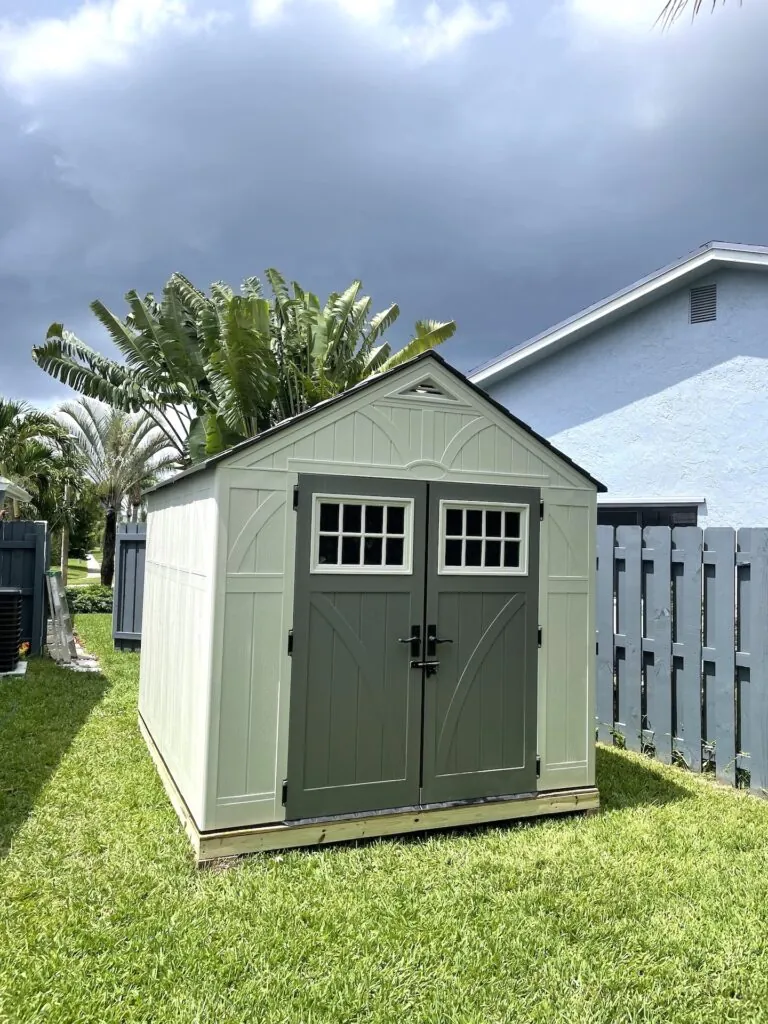 Green resin garden shed with windowed double doors beside a white fence