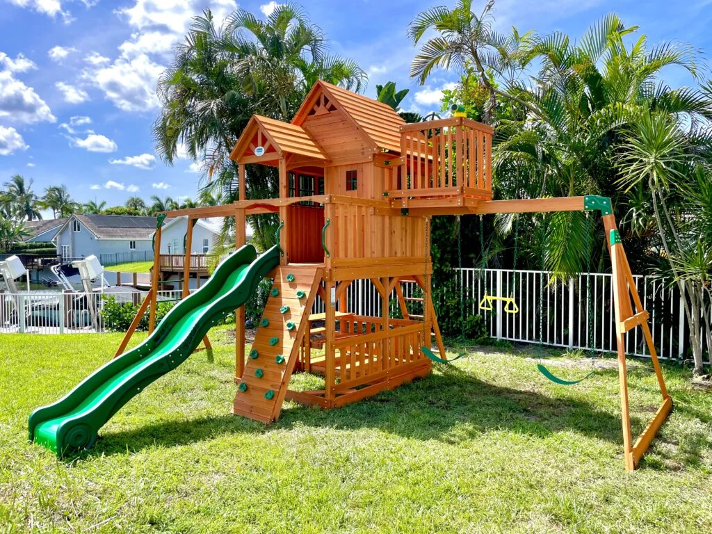 A finished wooden playset with swings and a slide in a sunny South Florida backyard, assembled by David Mertl