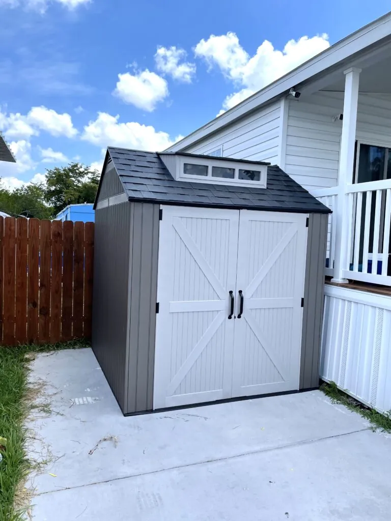 Light gray resin storage shed with double doors on a paved patio beside a house