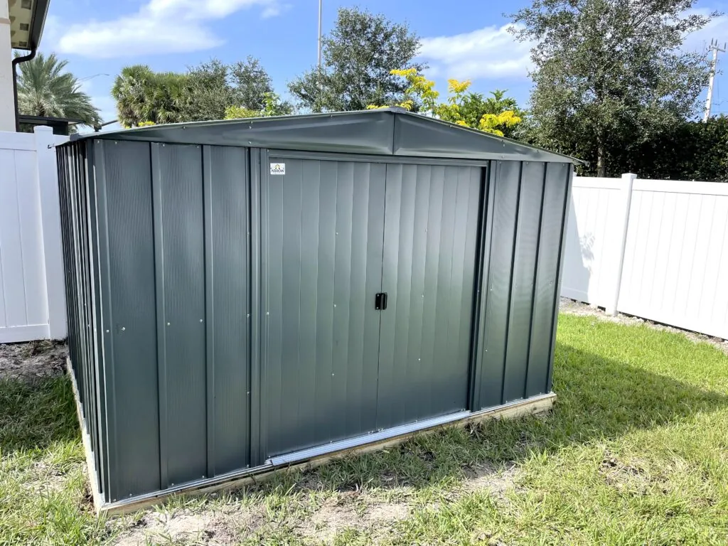 Wide gray metal storage shed with sliding doors beside a white backyard fence