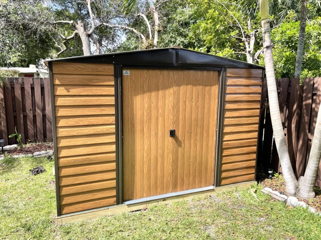 Wood-look storage shed with dark trim and double doors beneath backyard trees