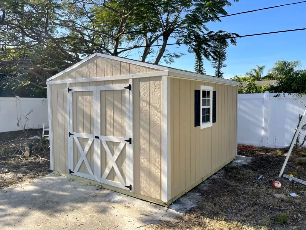 Beige storage shed with barn-style double doors and a side window in a fenced backyard